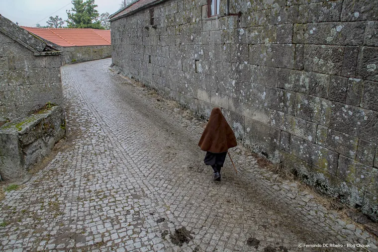 Rua de aldeia em calçada de granito, com uma pessoa de capa castanha a caminhar